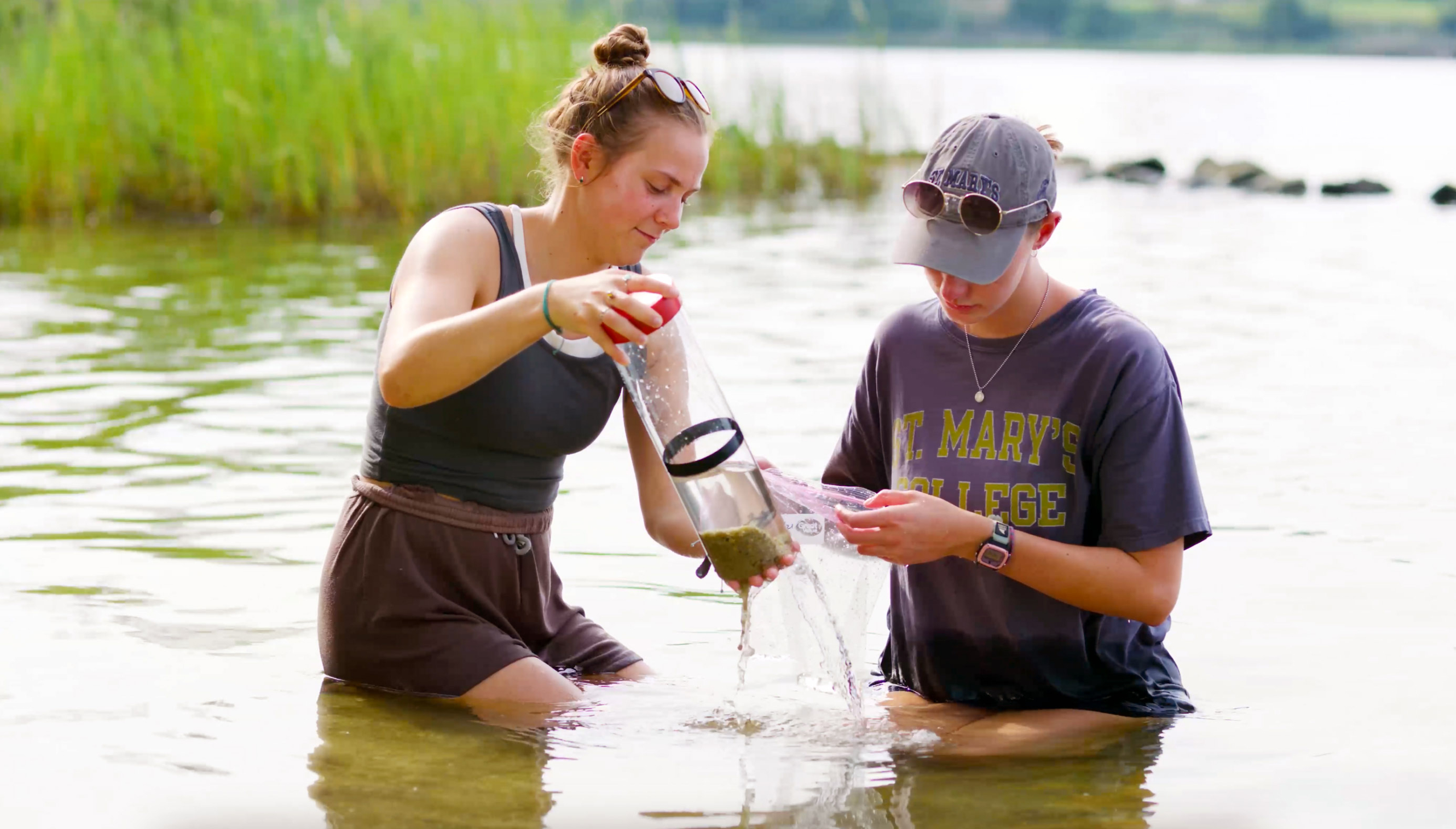 Two people kneel in shallow water, sampling; one pours water while the other examines it, with greenery and a shoreline in the background.